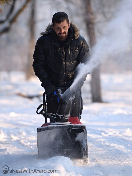 A man happily uses his snow blower while using a thick jacket