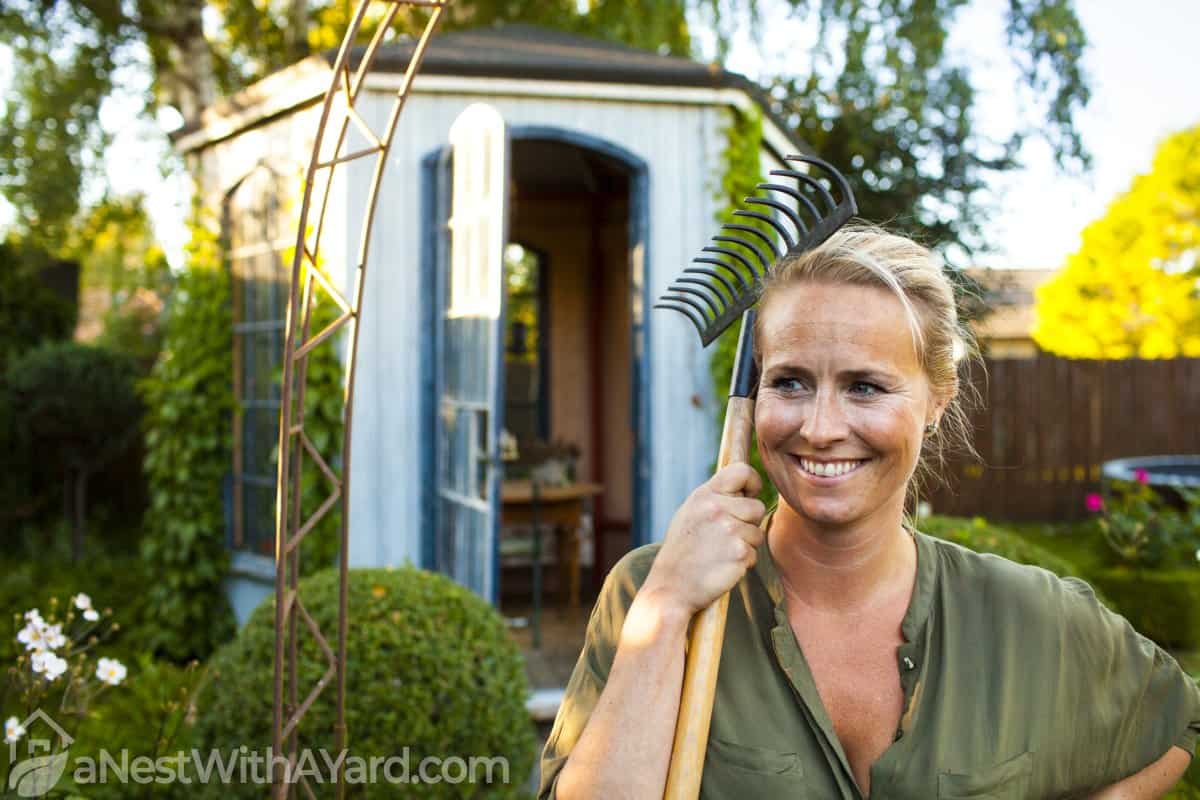 A woman standing near a shed holding a gardening tool in the hand
