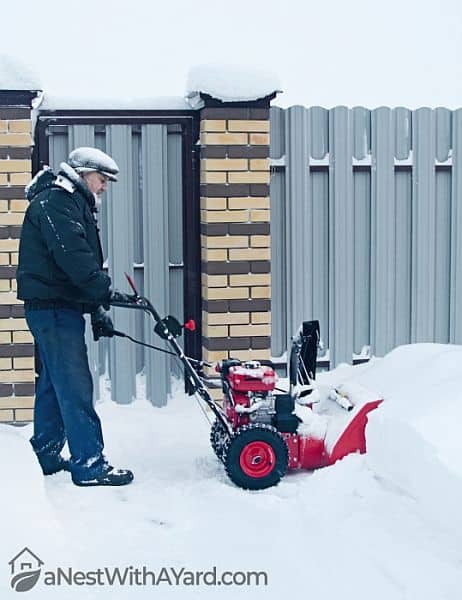 An old guy operating a red snow blower by his fence